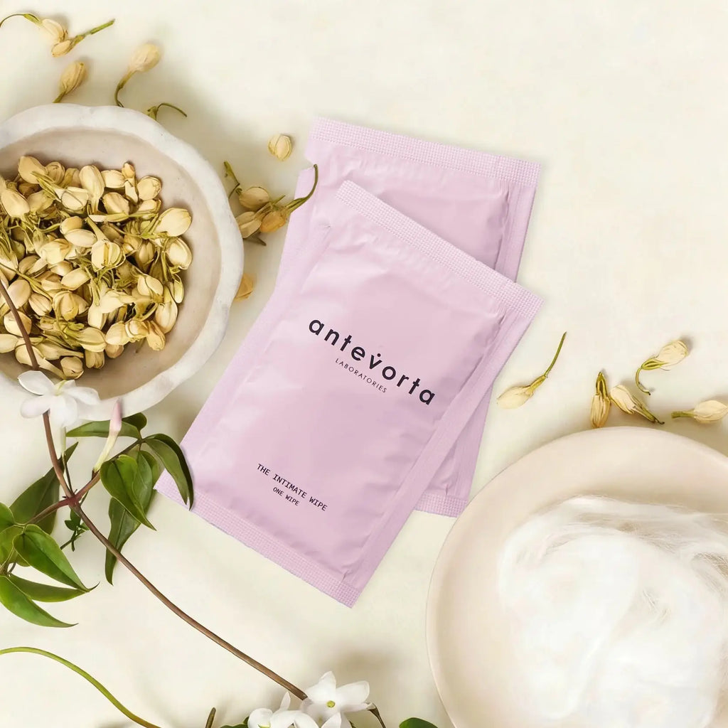Two lavender Antivortia skincare packets are arranged on a light cream surface with botanical props, including a bowl of dried jasmine flowers, green leaves, and a plate with white cotton pads.