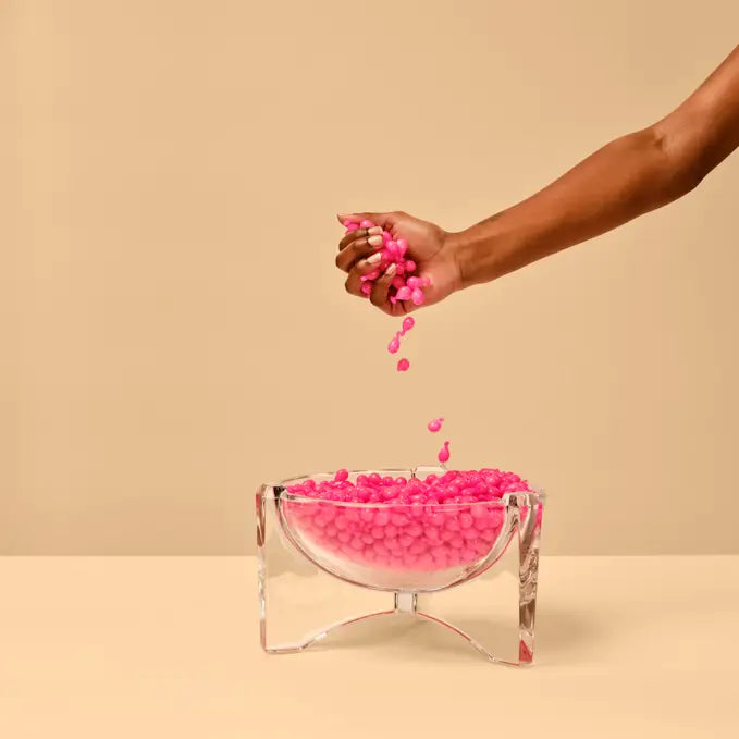 A hand holding a handful of bright pink candy above a clear glass bowl filled with pink candy, set against a warm beige background.