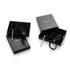 Two black Crave jewelry gift boxes shown open and closed on a white background, each containing a black leather bracelet and a slim metal necklace in different finishes.