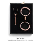 A jewelry mini set in pink and rose gold, displayed inside a black presentation box. The set includes a rose gold vesper pendant necklace, a pink ID cuff, and two rose gold ID cuffs.
