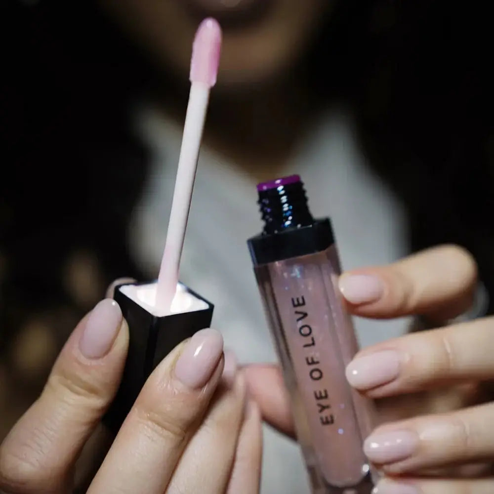 A close-up of a person holding a clear tube of lip gloss with a pink doe-foot applicator, showing the pink gloss inside. The tube has 'EYE OF LOVE' printed vertically on the front.