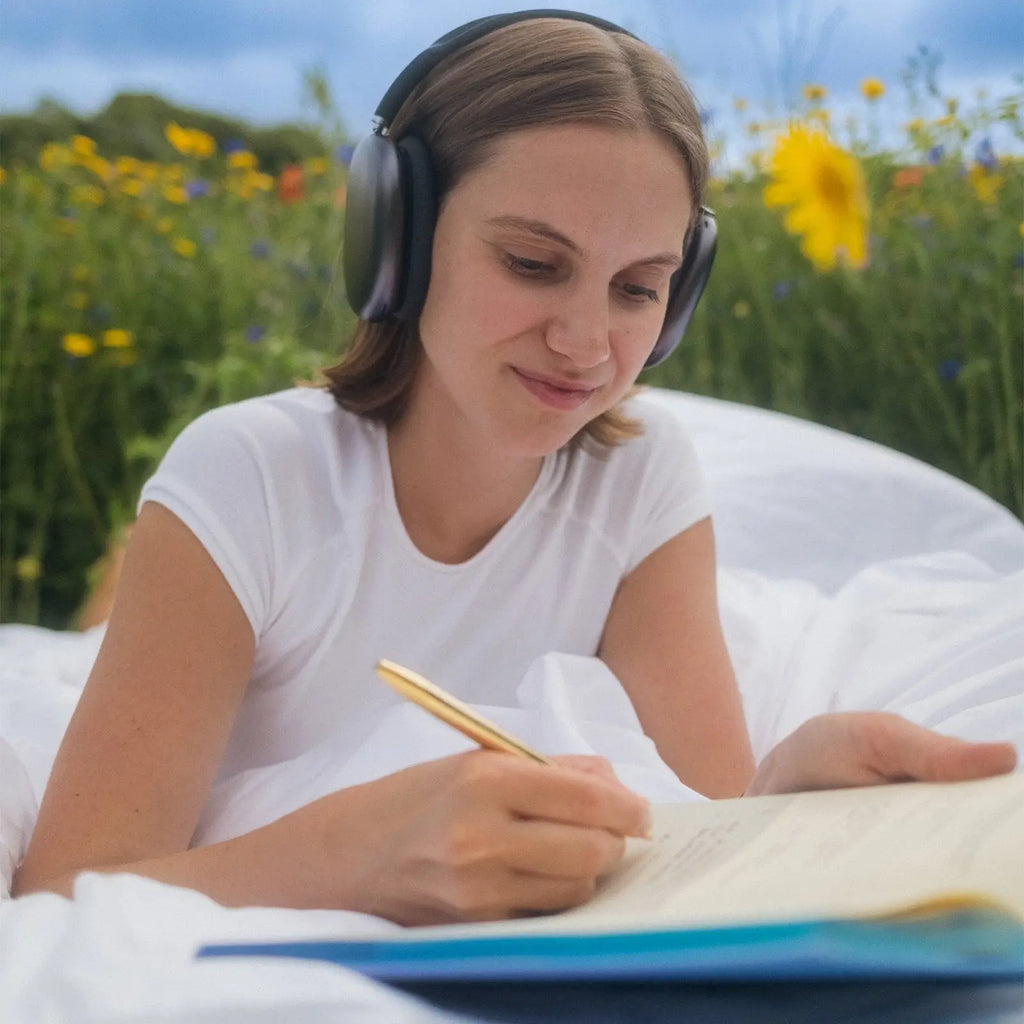 Woman wearing headphones and writing in a notebook outdoors with flowers and sky in the background