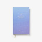 A hardcover journal with a blue-to-purple gradient cover, featuring gold foil text that reads 'The FIVE MINUTE JOURNAL' and the brand name 'Cala' near the bottom. A gold ribbon bookmark is visible at the bottom edge, set against a clean white background.