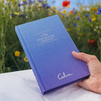 Blue journal titled 'The Five Minute Journal' held by a hand with a blurred natural background of flowers and sky.