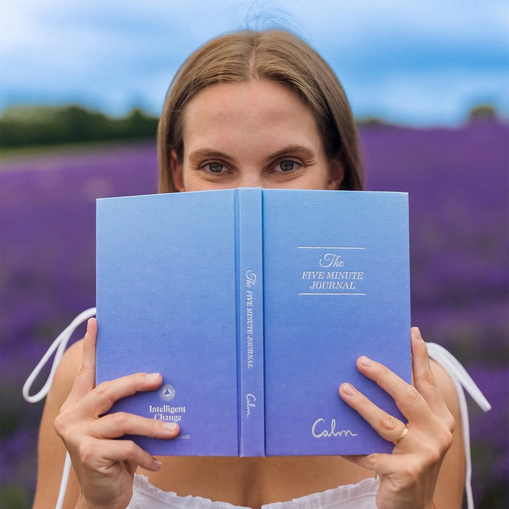 Woman holding a blue journal in front of her face with a lavender field in the background