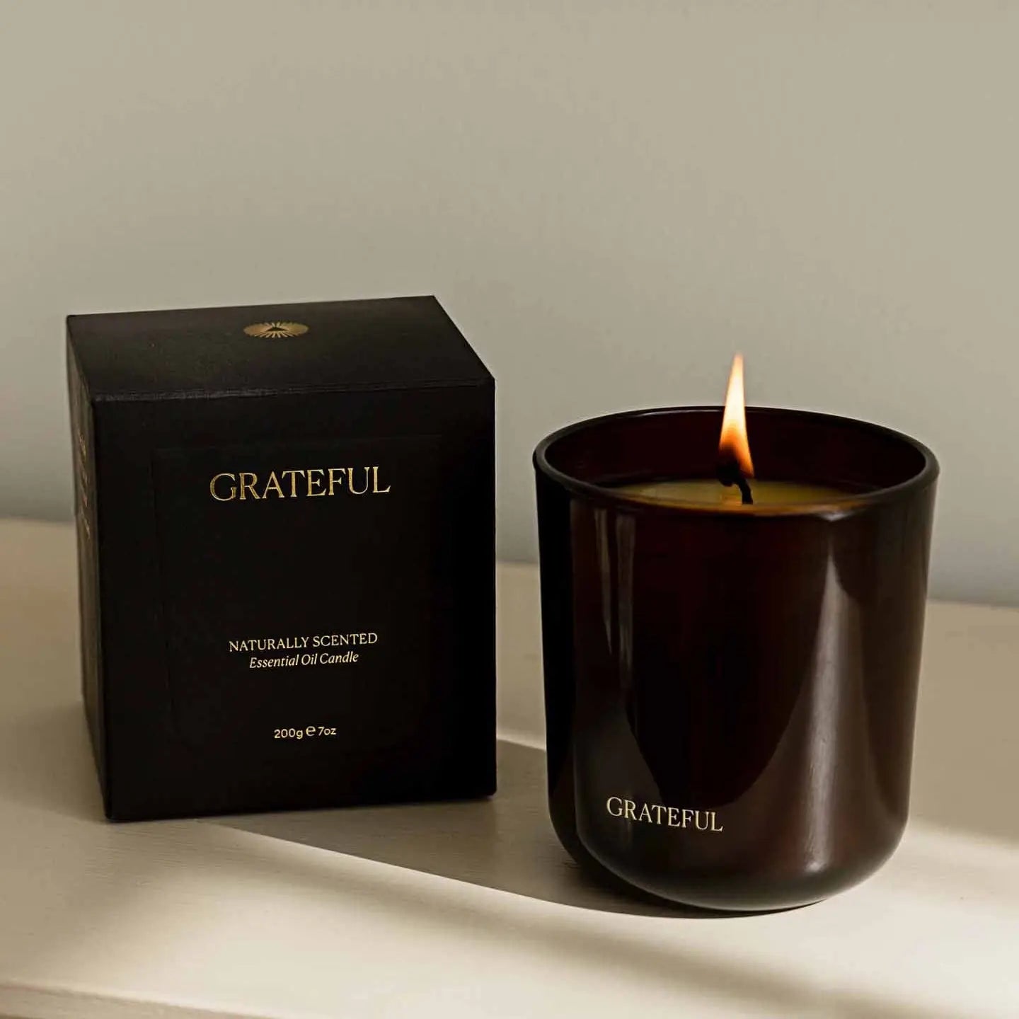 Black 'Grateful' candle with a lit wick next to its packaging on a light surface.