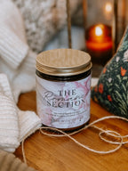 Candle labeled 'The Romance Section' on a wooden surface with soft lighting